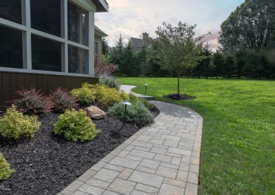 A stone walkway by Solid Brick Contracting curves past elegant landscaping and a screened porch, leading toward a grassy yard at sunset.