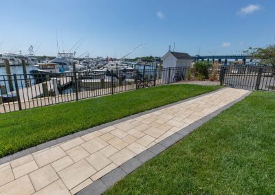 A paved walkway with lush green grass leads to a marina with boats docked and a small white shed by the water, showcasing quality landscaping in Aberdeen by Solid Brick Contracting.