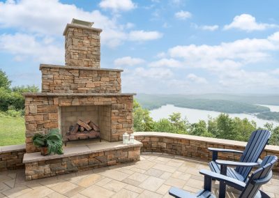Outdoor stone fireplace on a patio with blue chairs, overlooking a scenic lake and green hills—perfect for enjoying Aberdeen's tranquil beauty and expertly crafted hardscaping.