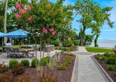 Paved walkway with hardscaping by landscaped garden in Aberdeen, MD, patio seating with umbrella, and a view of trees and water in the background.