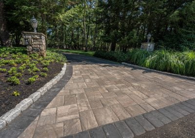 A stone driveway in Aberdeen, MD, leads to a tree-lined road with stone pillars and garden landscaping on both sides, crafted by Solid Brick Contracting.