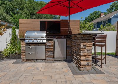 Outdoor kitchen in Aberdeen, MD featuring a stainless steel grill, fridge, TV, bar stools, and a red umbrella—perfect for entertaining on your hardscaping patio.