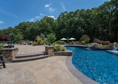 Spacious patio with expert hardscaping and stone pavers, a pool, umbrellas, and lush trees in the background under a blue MD sky.
