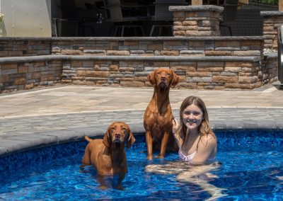 A girl in a pool with two brown dogs standing beside her, all looking at the camera, shows off beautiful landscaping in MD.