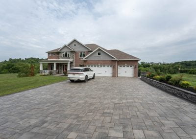 Spacious brick house in Aberdeen, MD with a three-car garage, white SUV on a wide stone hardscaping driveway, surrounded by lush green lawn and trees.