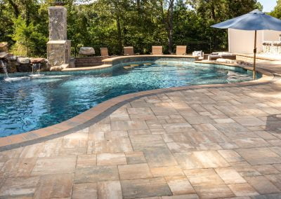 Curved swimming pool with stone patio, lounge chairs, umbrella, and lush landscaping in the background on a sunny day in Aberdeen, MD.