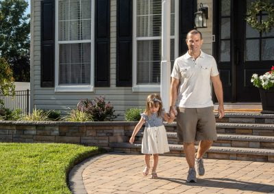 A man and a young girl holding hands, walking outside a house with steps and beautiful landscaping, possibly crafted by Solid Brick Contracting in MD.