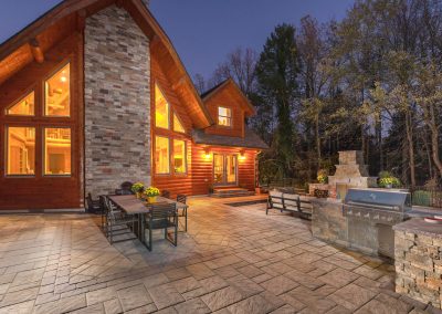 Large wooden house with bright windows, spacious hardscaping stone patio by Solid Brick Contracting, outdoor dining area, grill, and fireplace at dusk.