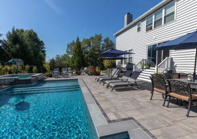 Backyard pool with lounge chairs, patio table, umbrellas, and a white house on a sunny day—expert hardscaping by Solid Brick Contracting brings this Aberdeen retreat to life.