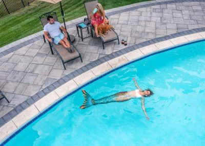 A woman in a mermaid tail floats in a pool surrounded by beautiful landscaping as two people relax on lounge chairs nearby.