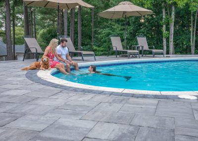 A couple with their dog enjoys the MD sunshine by a stylish, hardscaping-lined pool as a person in a mermaid tail swims nearby—an outdoor retreat crafted by Solid Brick Contracting.