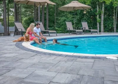 A couple with their dog enjoys the MD sunshine by a stylish, hardscaping-lined pool as a person in a mermaid tail swims nearby—an outdoor retreat crafted by Solid Brick Contracting.