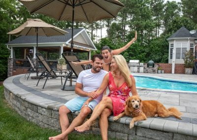 A couple sits by a beautifully landscaped pool with their dog, while a smiling woman poses excitedly behind them—showcasing the expert work of Solid Brick Contracting in MD.