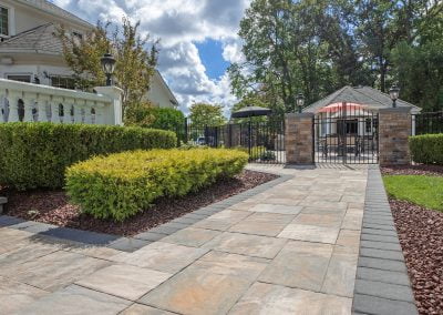 Sunny walkway with large stone pavers leads to a fenced area, bordered by green bushes and a white house—expertly crafted by Solid Brick Contracting in Aberdeen, MD.