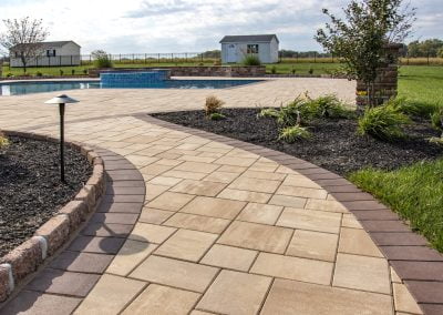 Curved stone walkway leading to a pool, surrounded by expert landscaping and lush grass, with a small shed in the distance. Designed and crafted by Solid Brick Contracting.