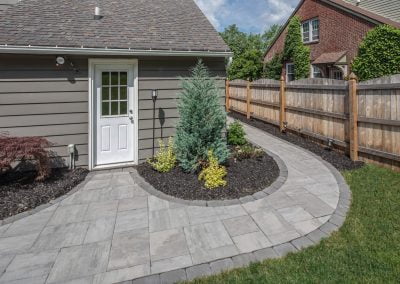 Curved stone walkway beside house, bordered by mulch, shrubs, and a wooden fence on a sunny day—showcasing expert hardscaping and landscaping by Solid Brick Contracting.