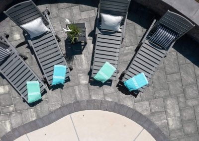 Five gray lounge chairs with pillows and teal towels sit on a stone patio by Solid Brick Contracting, showcasing expert landscaping, viewed from above.
