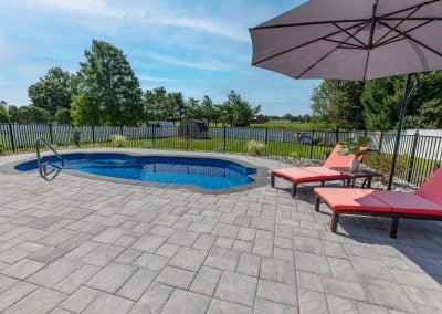 Charming small backyard pool with steps, two pink lounge chairs, umbrella, and beautiful landscaping. Expertly crafted by Solid Brick Contracting, MD—perfect for relaxing in your private oasis surrounded by trees and a fence.