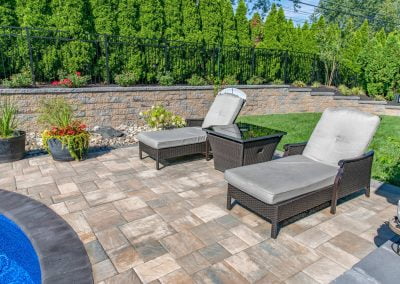 Two cushioned lounge chairs and a table on a stone patio beside a pool, surrounded by lush landscaping and a green hedge—expertly crafted by Solid Brick Contracting in MD.