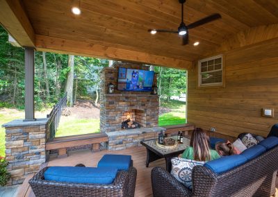 Two people relax on a covered patio by Solid Brick Contracting, watching TV near a fireplace and enjoying expert hardscaping, all surrounded by the peaceful woods in MD.