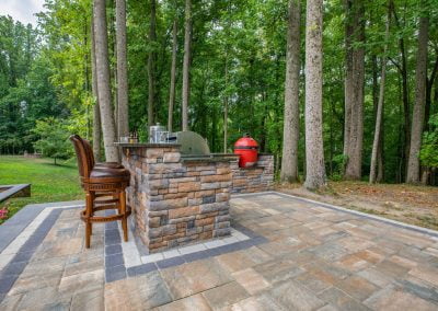 Outdoor patio with stone bar, barstools, and a red grill set among tall trees in a wooded area—expert hardscaping by Solid Brick Contracting creates a stylish Aberdeen retreat.