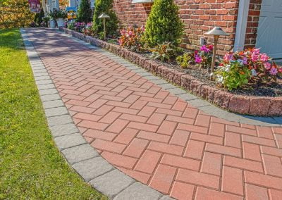Red brick walkway with hardscaping elements and a gray border, lined with flowers, bushes, and outdoor lights next to a brick house in Aberdeen, MD.