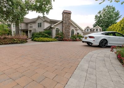 Large house with stone chimney, beautiful landscaping, garden, and a white car parked on a paved driveway under a partly cloudy MD sky.