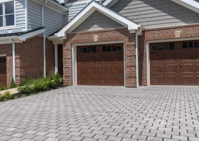 Brick house in Aberdeen, MD, with double wooden garage doors and a wide paved driveway, built by Solid Brick Contracting under a blue sky with clouds.