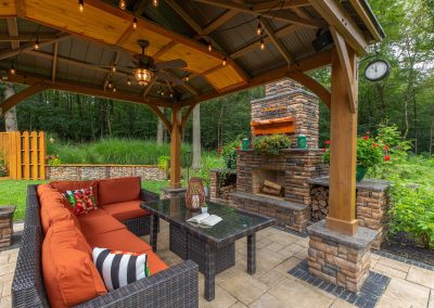 A cozy patio by Solid Brick Contracting features orange seating, a glass table, string lights, and a stone fireplace under a wooden pergola—perfect for enjoying expert hardscaping and inviting outdoor living.