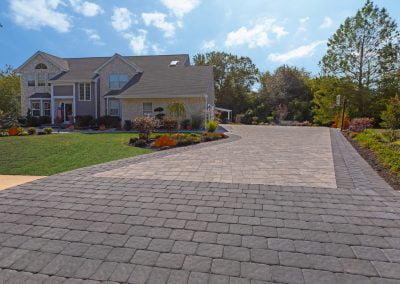 A large MD house with beautiful landscaping, a landscaped yard, and a wide, patterned paver driveway under a blue sky.