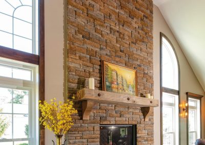 Living room with tall stone fireplace, wood mantle, large windows, a vase with yellow flowers, and views of expertly designed landscaping by Solid Brick Contracting.