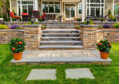 Stone steps by Solid Brick Contracting lead to a patio with red chairs, potted flowers, and neatly landscaped garden beds—a perfect example of expert hardscaping in MD.