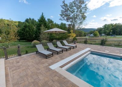 Four lounge chairs with an umbrella beside a pool, surrounded by lush trees and a scenic outdoor landscape, showcase expert landscaping and hardscaping by Solid Brick Contracting.