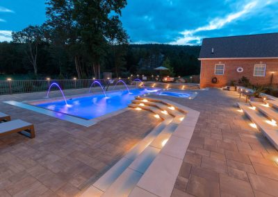 Modern pool with glowing water jets, surrounded by lit steps and lounge chairs at dusk—expert landscaping and hardscaping by Solid Brick Contracting complete the stunning scene.