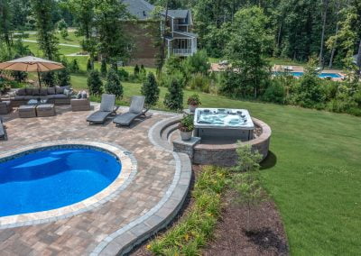 A backyard in MD featuring a pool, hot tub, lounge chairs, patio seating, and green lawn bordered by trees—perfectly enhanced with expert hardscaping by Solid Brick Contracting.