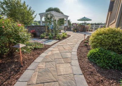 Curved stone walkway features expert hardscaping through landscaped garden, leading to a pergola and outdoor seating area in sunny Aberdeen.