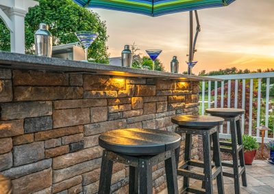 Chic outdoor stone bar with stools, martini glasses, and an umbrella at sunset showcases elegant hardscaping, overlooking a garden in Aberdeen, MD.