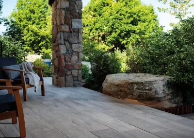 Wood-look tile patio with stone pillar, wooden chairs, and lush green trees and bushes—expert hardscaping and landscaping by Solid Brick Contracting set the perfect outdoor scene.