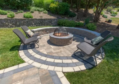 Three wicker chairs surround a stone fire pit on a circular stone patio, creating an inviting backyard retreat in Aberdeen, MD landscaping.