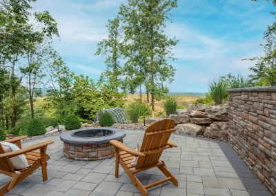 Two wooden chairs face a stone fire pit on a paved patio, expertly crafted by Solid Brick Contracting in Aberdeen, MD, with trees and blue sky in the background.