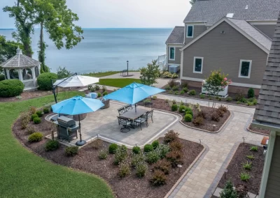 Backyard patio by Solid Brick Contracting features blue umbrellas, dining tables, a grill, gazebo, and stunning lake view—perfect landscaping for your MD home behind a tan house.