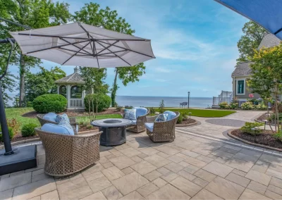 Charming patio with wicker chairs, umbrella, and fire pit overlooking a lake in Aberdeen, MD. A gazebo and trees set the scene—beautifully crafted by Solid Brick Contracting.