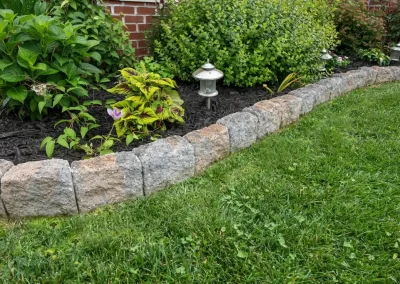A stone border showcases expert hardscaping, separating a mulched garden with plants and lanterns from a grassy lawn—perfect for elevating Aberdeen landscaping.