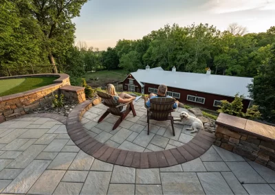 A couple and their dog relax on patio chairs overlooking a scenic yard and a large barn at sunset in Aberdeen, MD, crafted by Solid Brick Contracting.