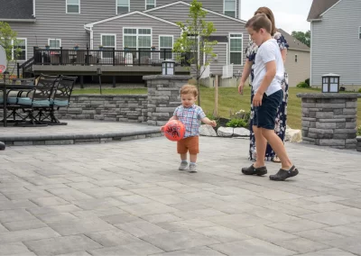 A toddler holds a red ball while walking outside with a woman and an older boy on a hardscaping patio.
