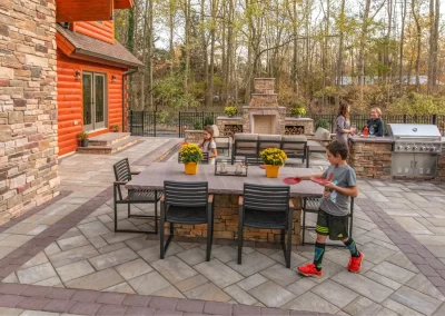 Children and adults enjoy a backyard patio with a table, fireplace, and grill surrounded by trees, featuring beautiful landscaping from Solid Brick Contracting.