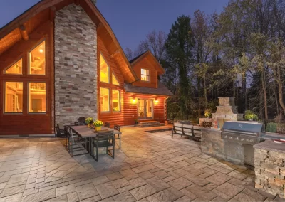 Modern log cabin in Aberdeen, MD with large windows, lit patio, outdoor dining area, grill, and fireplace at dusk—crafted by Solid Brick Contracting.