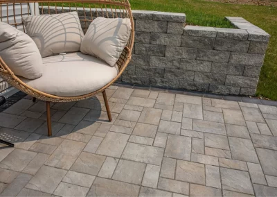 Wicker chair with cream cushions on a stone patio, expertly crafted by Solid Brick Contracting, beside a low stone wall and grass—perfect for Aberdeen-inspired hardscaping.