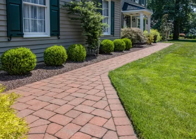 Curved brick walkway by Solid Brick Contracting in Aberdeen, MD, with shrubs lining the house, bordered by green grass and landscaped plants.
