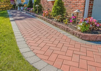 Brick walkway bordered with gray stones, lined with flowers, shrubs, and outdoor lights beside a brick house—expertly crafted by Solid Brick Contracting, your trusted MD landscaping specialists.